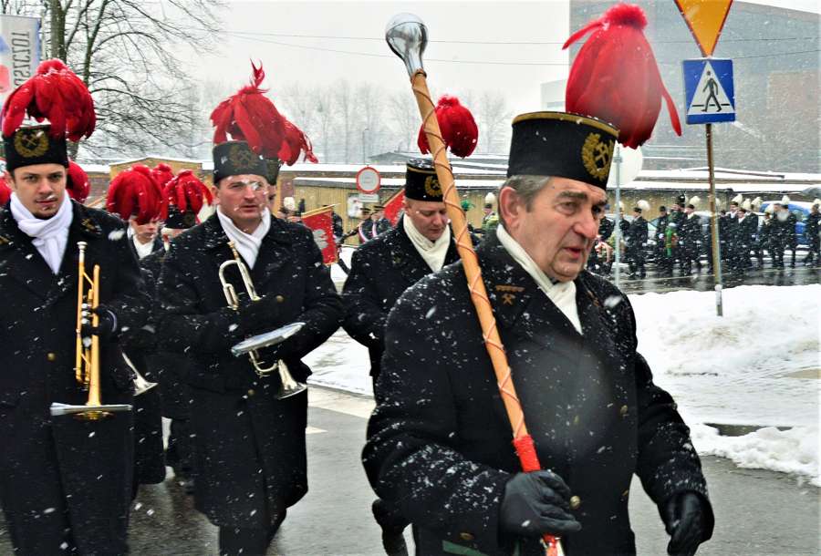 Miners in black gala coats and red-plumed czako hats march with brass instruments through falling snow in Piekary Śląskie