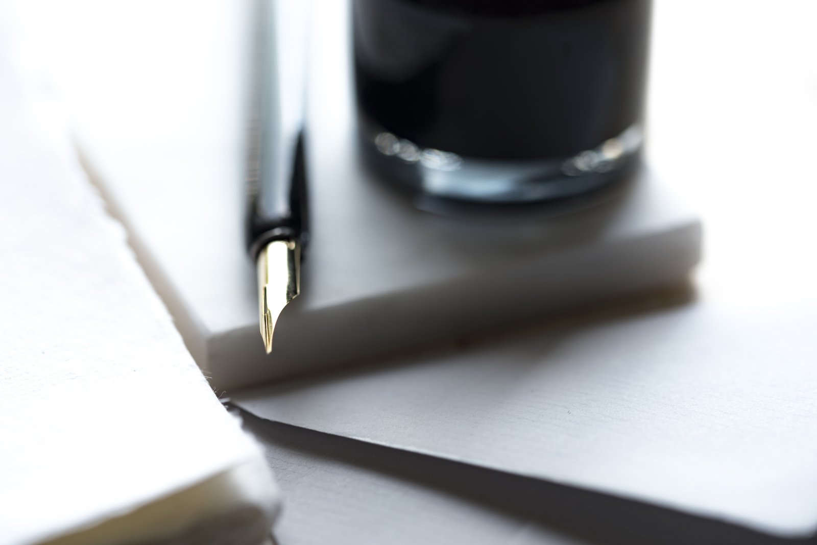 A close-up of a metal pen nib beside a dark ink container and pale paper