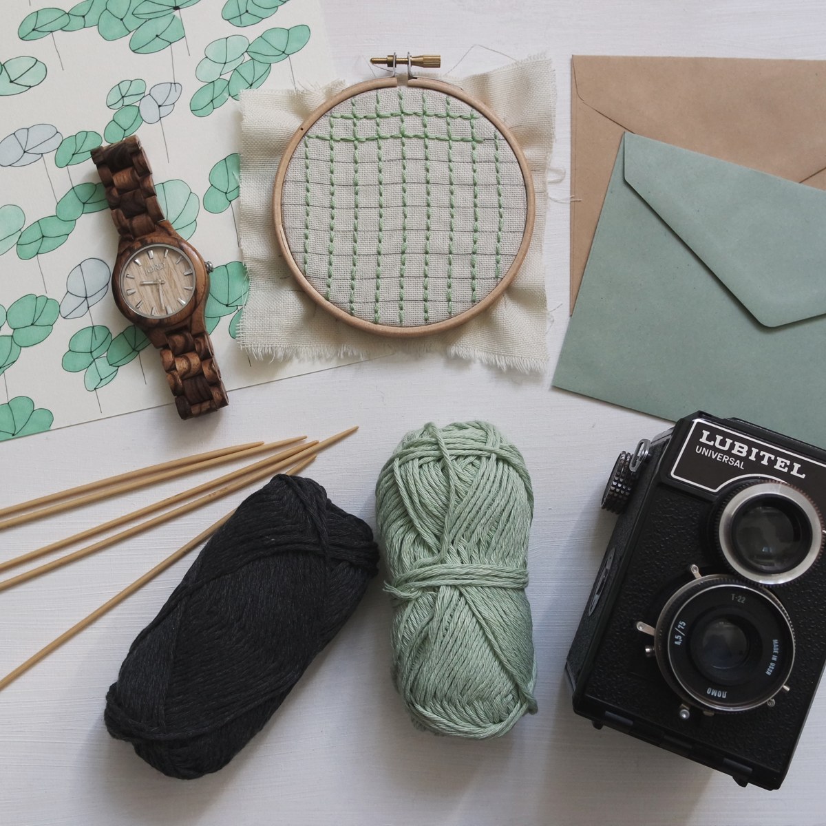 A wooden hoop holding pale fabric with green stitches, beside yarn, knitting needles, a watch and a camera
