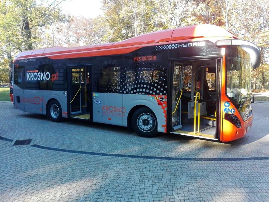 Red and black MKS Krosno hybrid bus parked on a paved square