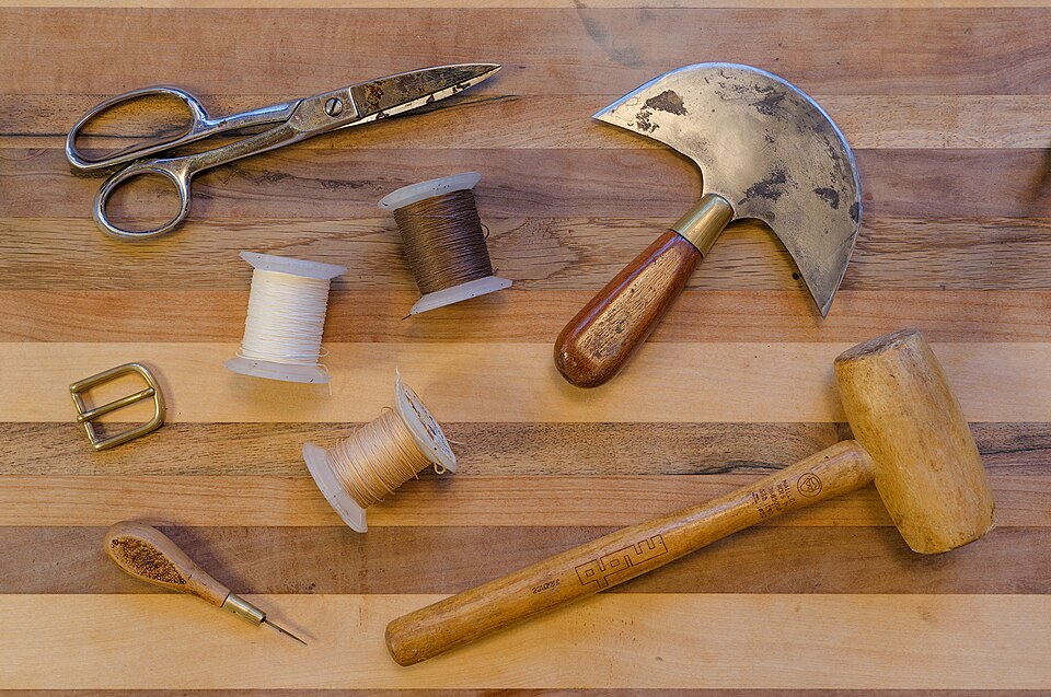 Leatherworking scissors, thread, a half-moon knife, awl, buckle and wooden mallet on a wooden surface