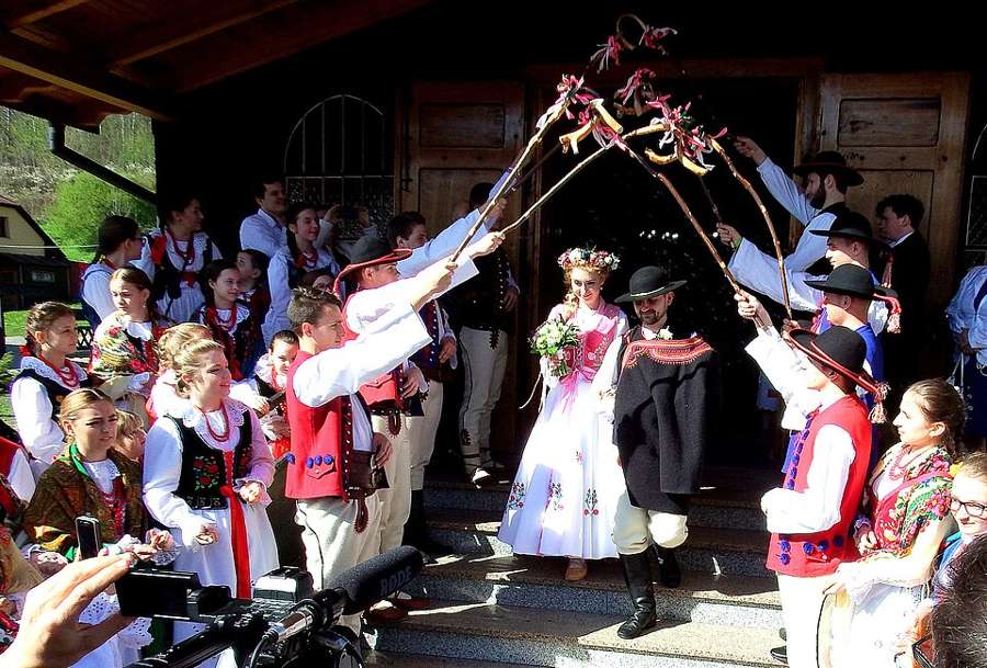 Highland wedding guests in folk costume welcome the newlyweds outside a wooden church in Juszczyna
