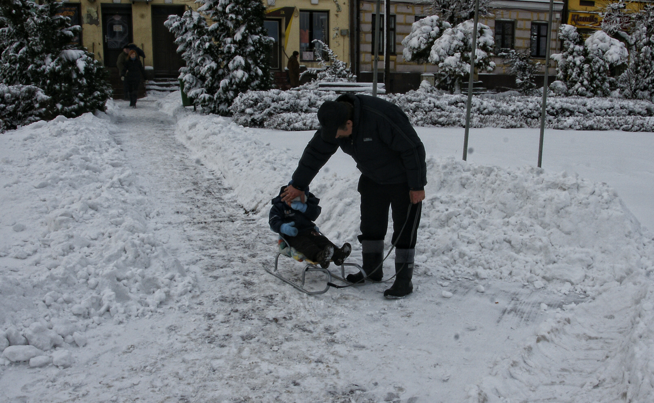 A parent pulling a small child on a sled along a snowy city path in Poland