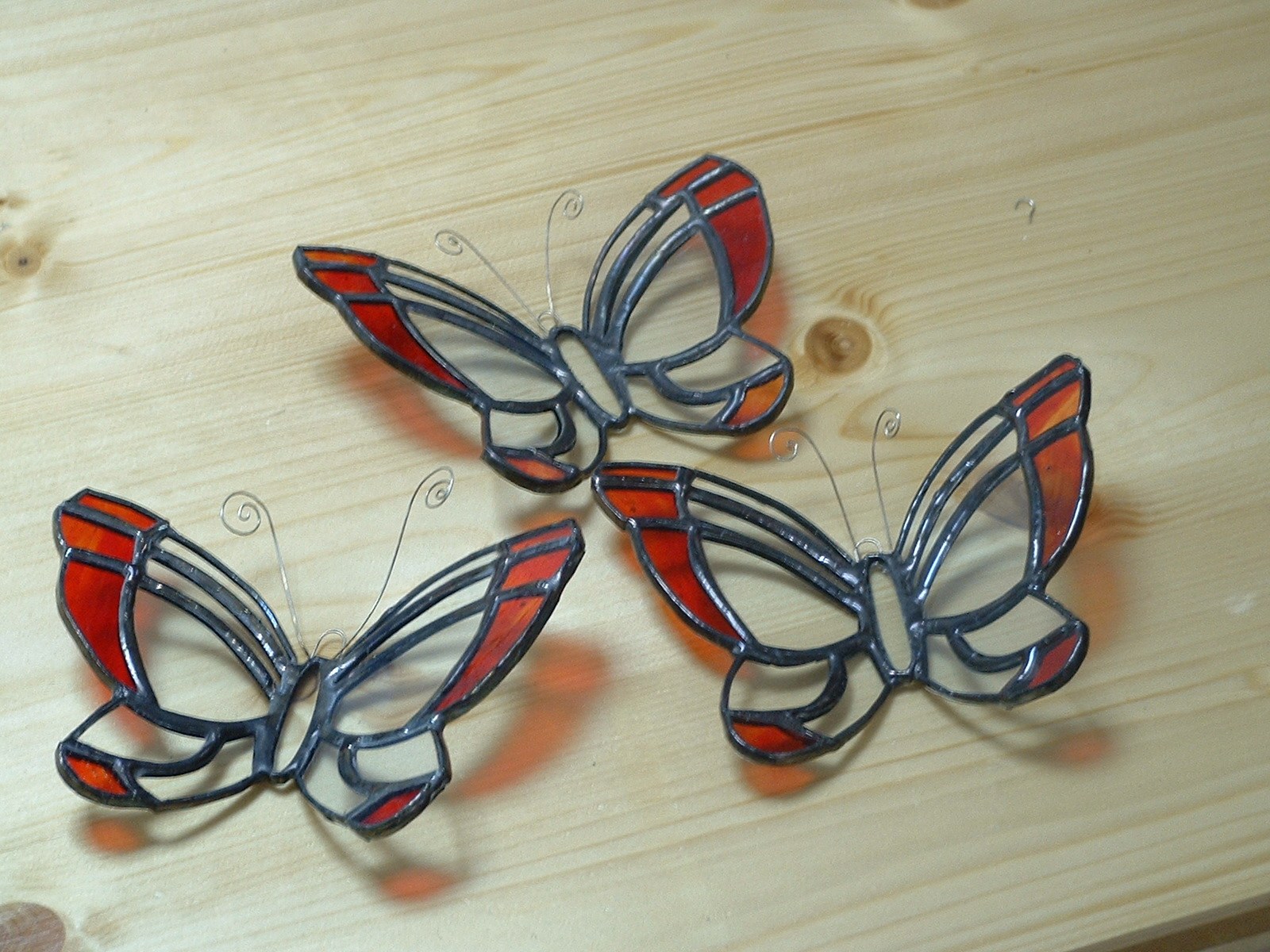 Three red and clear butterfly-shaped copper-foil glass ornaments with dark finished joints on a wooden surface