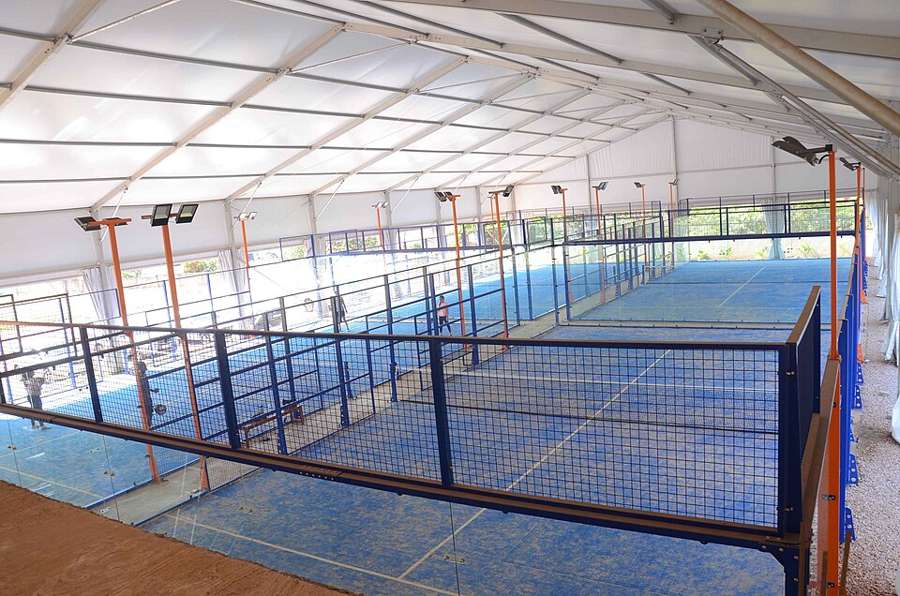 A row of blue indoor padel courts with glass walls under a white canopy roof